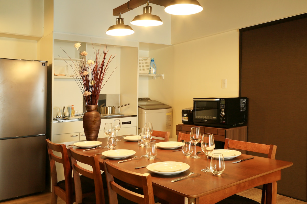 A dining area is presented with a wooden table set for six, featuring clear plates and glasses. Overhead, three pendant lights provide warm illumination. A refrigerator and microwave are positioned nearby, with a kitchen area visible in the background. Decorative elements include a vase of tall flowers.