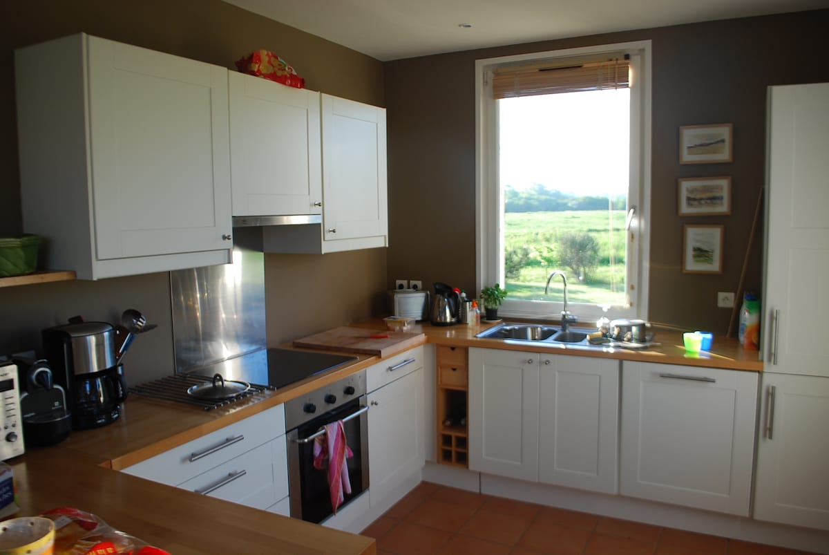 A modern kitchen features white cabinetry paired with warm wood countertops. Appliances include a stovetop, oven, and coffee maker. Natural light floods in through a large window, which overlooks greenery outside. Decorative elements, such as framed art, add character to the space.