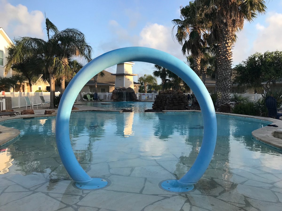 A circular blue archway frames the tranquil pool area, reflecting the surrounding palms in the water. The pool's surface is calm, with a decorative fountain visible in the background amidst a well-landscaped environment.