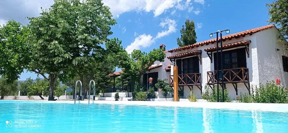 A serene outdoor scene features a clear, light blue pool framed by lush greenery and a few trees. The traditional-style home, with white walls and terracotta roofs, presents spacious balconies with wooden railings, offering a peaceful retreat surrounded by nature.