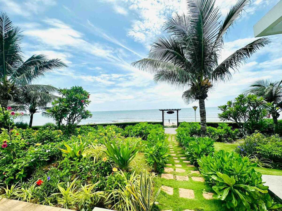 A well-maintained garden featuring lush greenery and vibrant flowers leads to a tranquil beachfront. Palms stand tall along the path, guiding the way towards the ocean. The sky above is filled with soft clouds, creating a serene atmosphere.