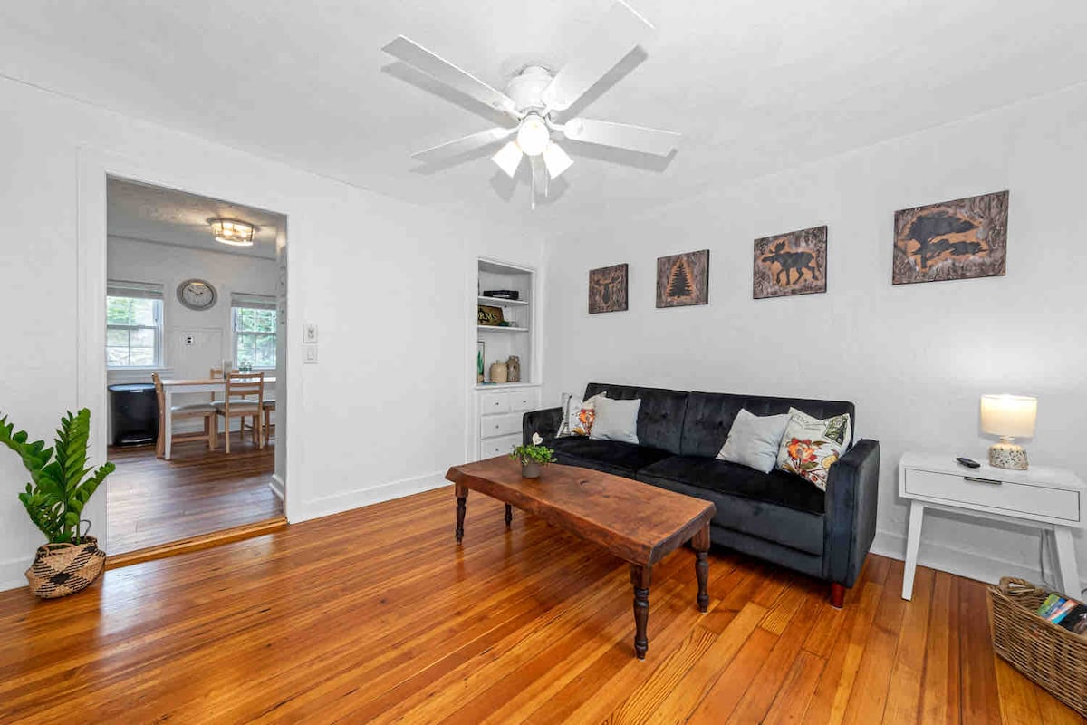 A spacious living room features a dark sofa adorned with decorative pillows, situated next to a wooden coffee table. Light wood flooring complements the neutral wall color. Nearby, a ceiling fan provides airflow, and artworks adorn the walls, enhancing the relaxed atmosphere.