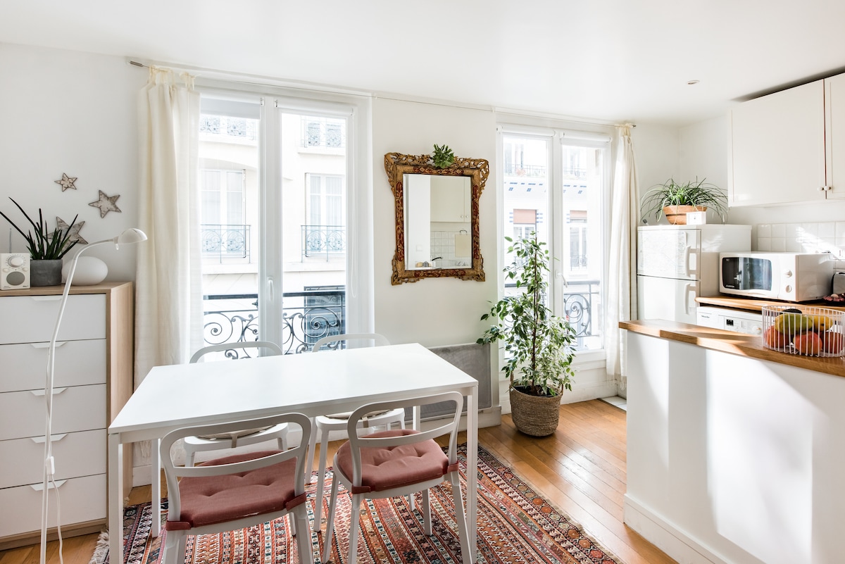A light-filled dining area features a white table surrounded by four chairs, with soft pink cushions. A large mirror is mounted on the wall, reflecting natural light. An indoor plant adds greenery, while a small white cabinet and a kitchenette are visible in the background.