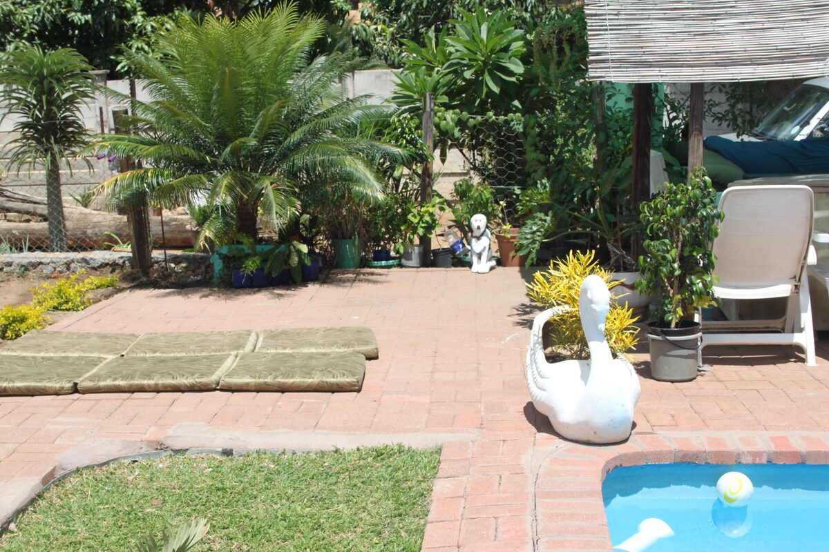 A garden area features neatly arranged green plants and a white swan sculpture beside a small swimming pool. Outdoor seating is provided near the pool, accompanied by a covered area. The ground is paved with red bricks, creating a tranquil and inviting environment.