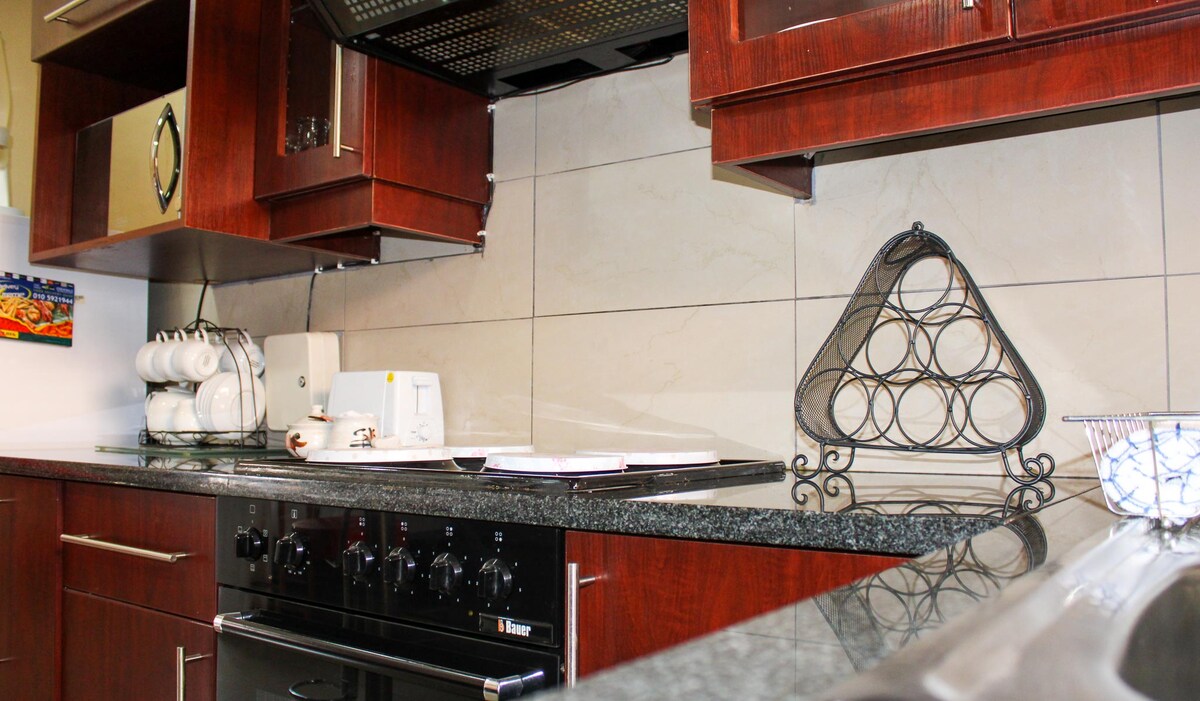 A modern kitchen is presented, featuring dark wood cabinetry and a dark granite countertop. Essential appliances include a stove, a microwave, and a coffee maker. A decorative wire basket adds a touch of elegance beside neatly arranged white dinnerware, enhancing the functional and inviting design.