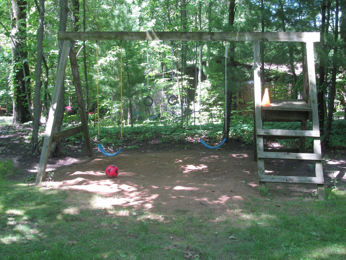 A wooden swing set is situated in a grassy area, surrounded by trees. Two swings are hanging from the frame, and a slide is attached at one end. A red soccer ball rests on the ground, with a traffic cone positioned nearby.