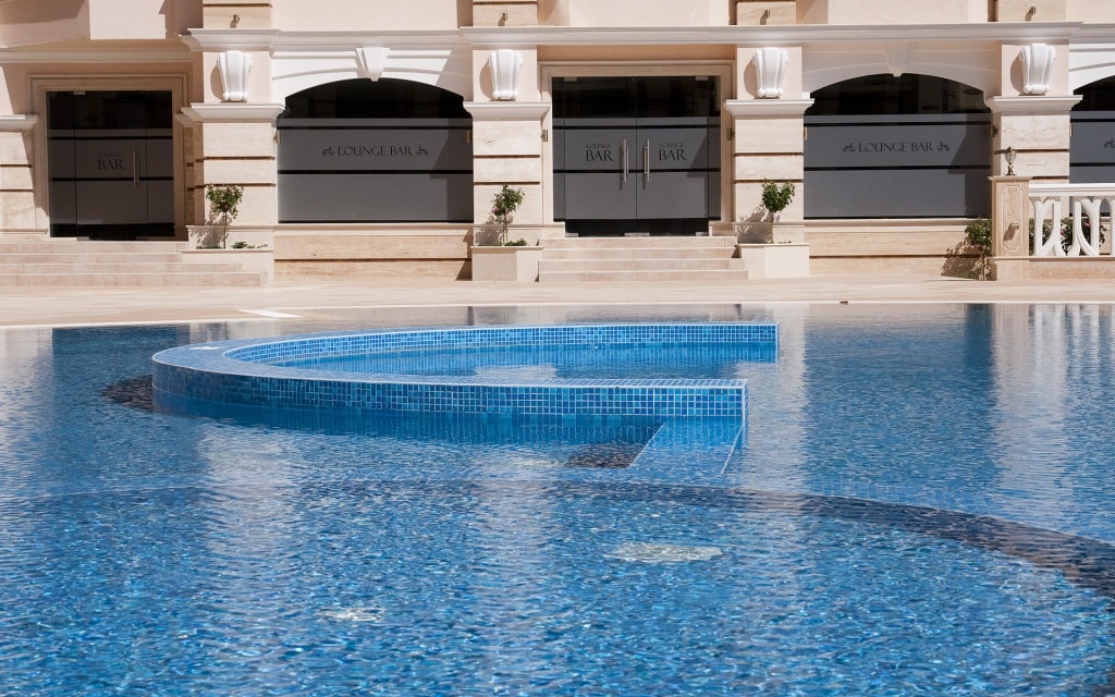 A large, inviting swimming pool is featured, with crystal-clear blue water shimmering under the sun. Its unique curved shape creates a modern aesthetic, while surrounding areas are well-maintained with potted plants and stylish architecture visible in the background.