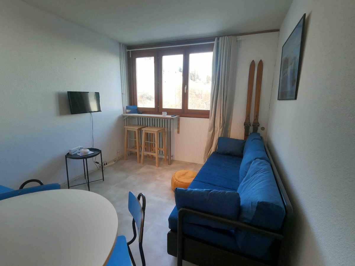 A bright living area is presented with a blue sofa and small round table. A mounted flat-screen TV is positioned on the wall opposite a window, allowing natural light to illuminate the space. Ski equipment is leaned against the wall, adding a touch of mountain charm.