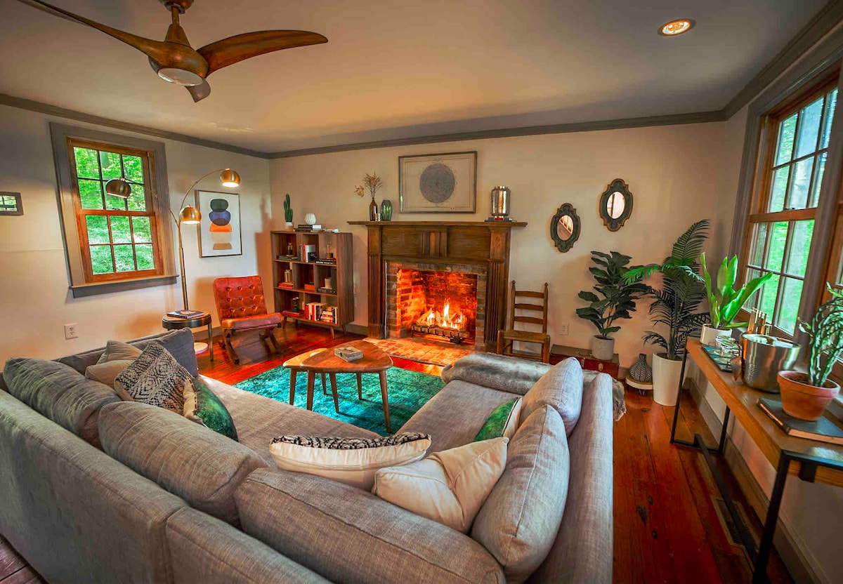 A cozy living area features a large sectional sofa arranged around a warm brick fireplace. Natural light streams through multiple windows, illuminating plants and artwork. Vintage armchairs provide additional seating, while a colorful area rug adds a touch of comfort to the hardwood floor.