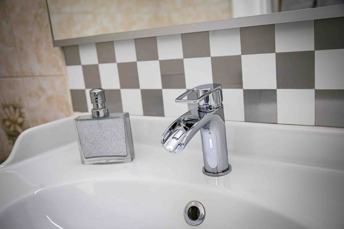 A clean and modern bathroom sink is displayed, featuring a sleek chrome faucet beside a glass soap dispenser. The backdrop consists of a checkered tile pattern, creating a subtle contrast with the smooth white sink surface.