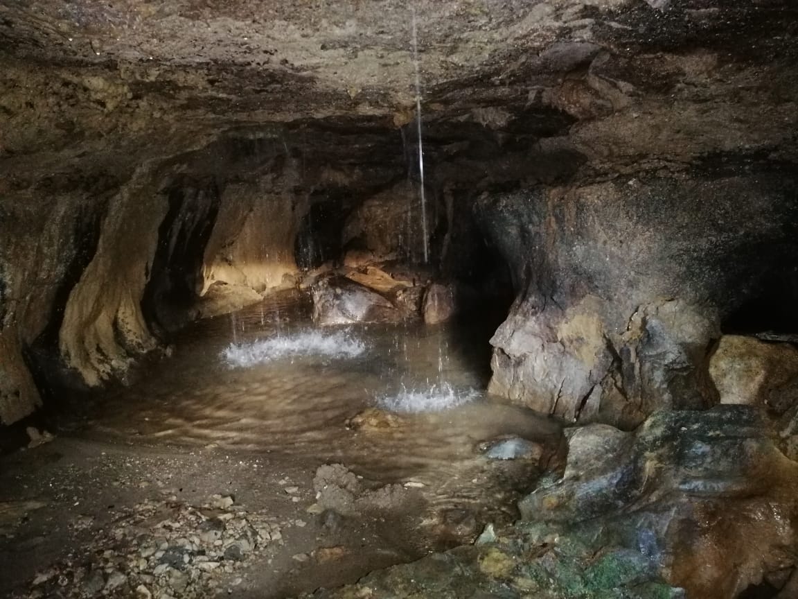 A cave interior is depicted, featuring rough stone walls and a small pool of clear water. Water droplets can be seen cascading from the ceiling, creating gentle ripples on the surface below. The lighting casts soft shadows, enhancing the natural textures of the rock formations.