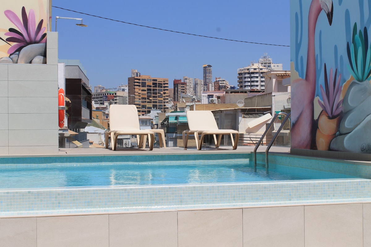 A rooftop pool surrounded by a modern deck features two lounge chairs positioned near the water. Vibrant murals depicting tropical scenes are visible on adjacent walls. The city skyline can be seen in the background under a clear blue sky.