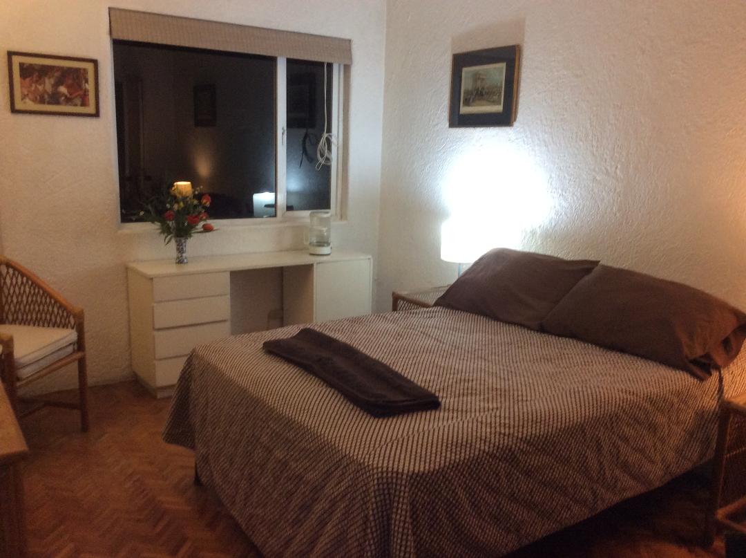 A spacious bedroom is illuminated by a soft bedside lamp, featuring a comfortable bed with a patterned coverlet. A desk is positioned near a window, with a vase of flowers and framed artwork adorning the walls, complementing the warm, inviting atmosphere.
