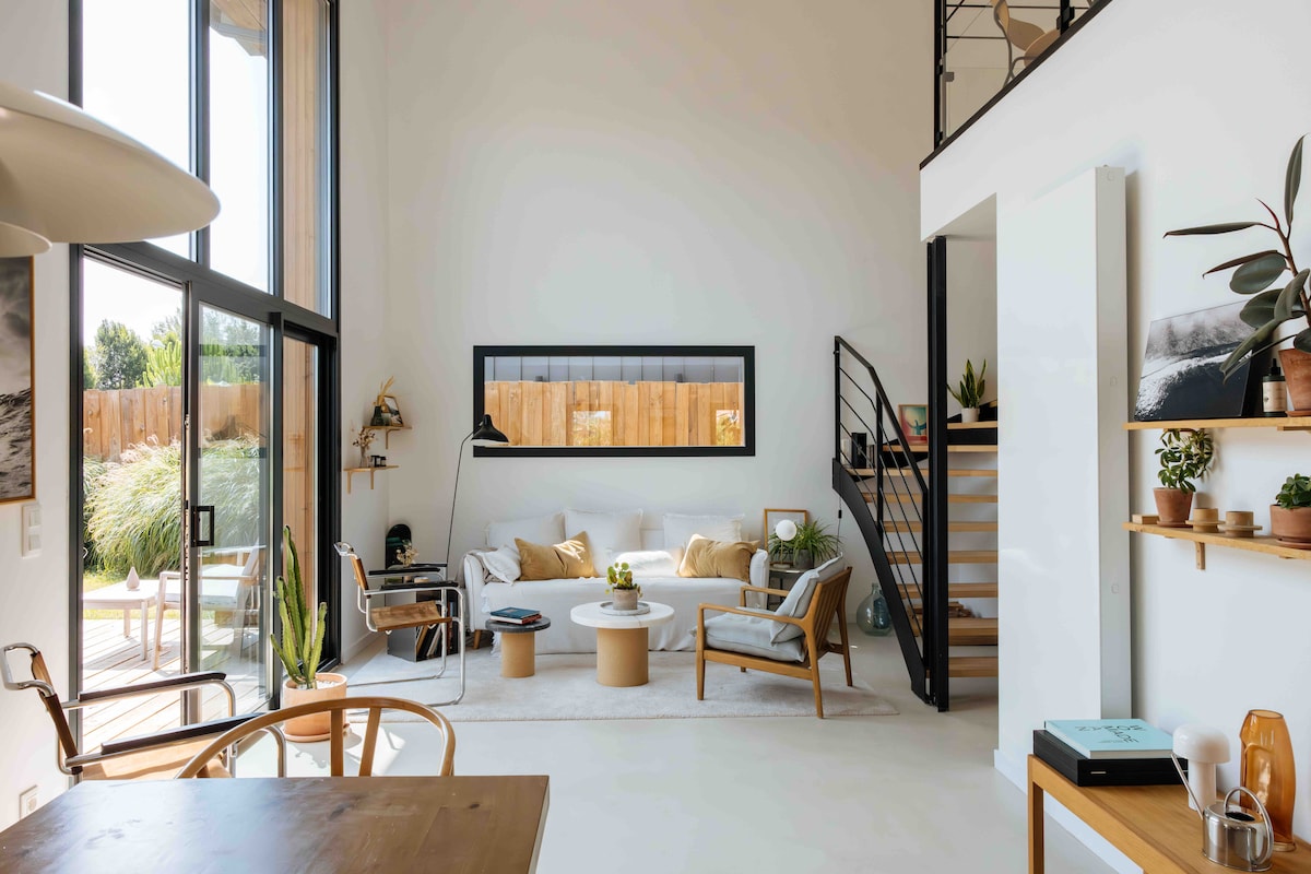 A bright living area features high ceilings with large windows, allowing natural light to fill the space. Comfortable seating is arranged around a round coffee table, with plants adorning shelves nearby. A spiral staircase leads to an upper level, seamlessly integrating the living space.