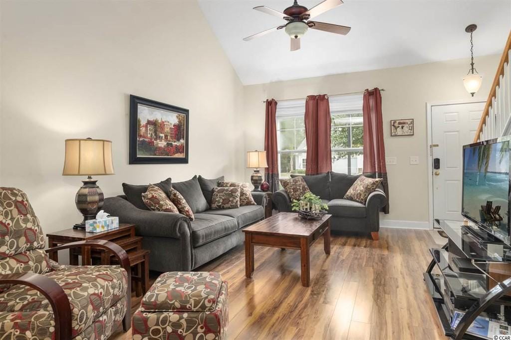 A cozy living area features plush seating with decorative cushions, complemented by a wooden coffee table. Natural light enters through large windows adorned with curtains. A ceiling fan circulates air, while a television is positioned on a glass console, enhancing the inviting atmosphere.