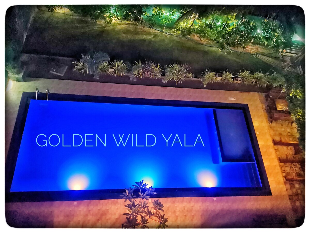 An aerial view of a modern swimming pool illuminated in shades of blue, set in a landscaped area. The pool features a sleek rectangular shape, with the name 'GOLDEN WILD YALA' displayed prominently across its surface. Surrounding greenery complements the tranquil setting.
