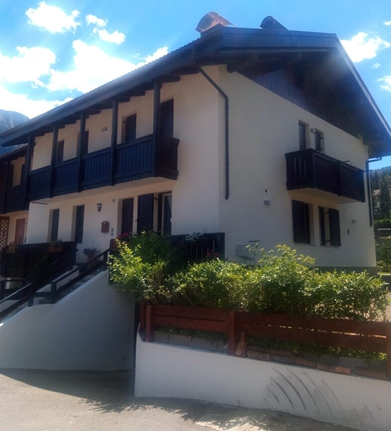 The apartment building features a two-story structure with a sloped roof and wooden balconies adorned with planters. An inviting entrance is complemented by a flight of stairs leading to the front door. Lush greenery frames the property, enhancing the welcoming appearance.
