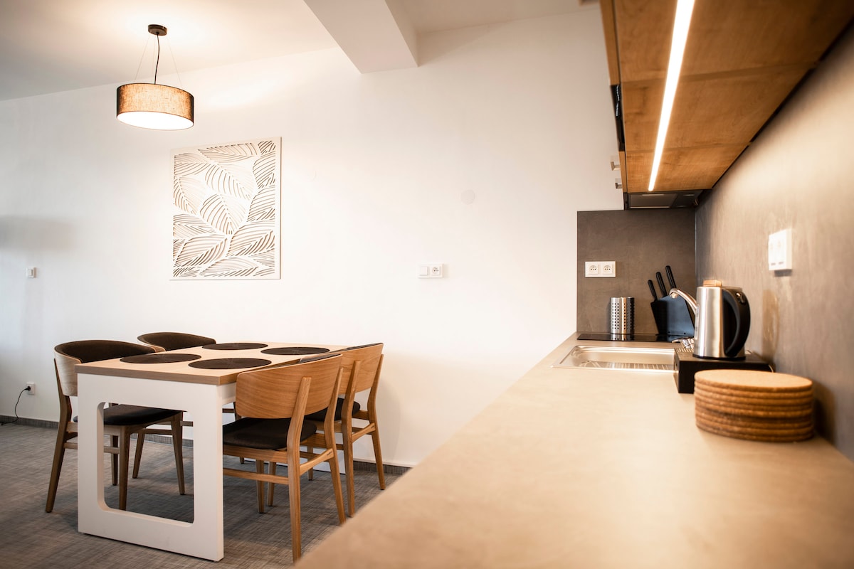 A modern dining area is designed with a sleek white table surrounded by four wooden chairs. A light fixture hangs above, while a minimalistic wall art piece adds a touch of decor. The adjacent kitchen features a countertop with appliances, promoting an inviting atmosphere.