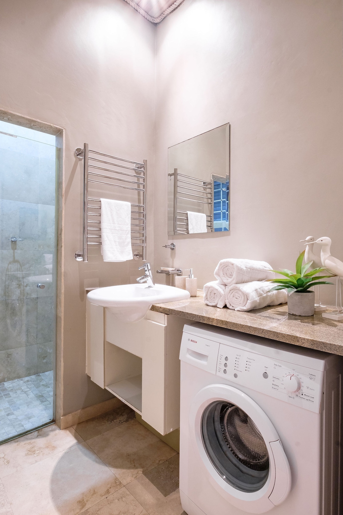 The bathroom features a compact design with a modern sink and a washing machine. Soft, rolled towels are neatly arranged next to a potted plant. A glass shower with textured tiles is visible, and light reflects gently off the mirrors, enhancing the space.