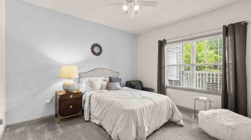 A bright bedroom features a spacious bed layered with a light-colored blanket and accent pillows. A bedside table with a lamp provides soft illumination, while a cozy chair sits in the corner next to a window adorned with curtains. A small table is placed nearby.