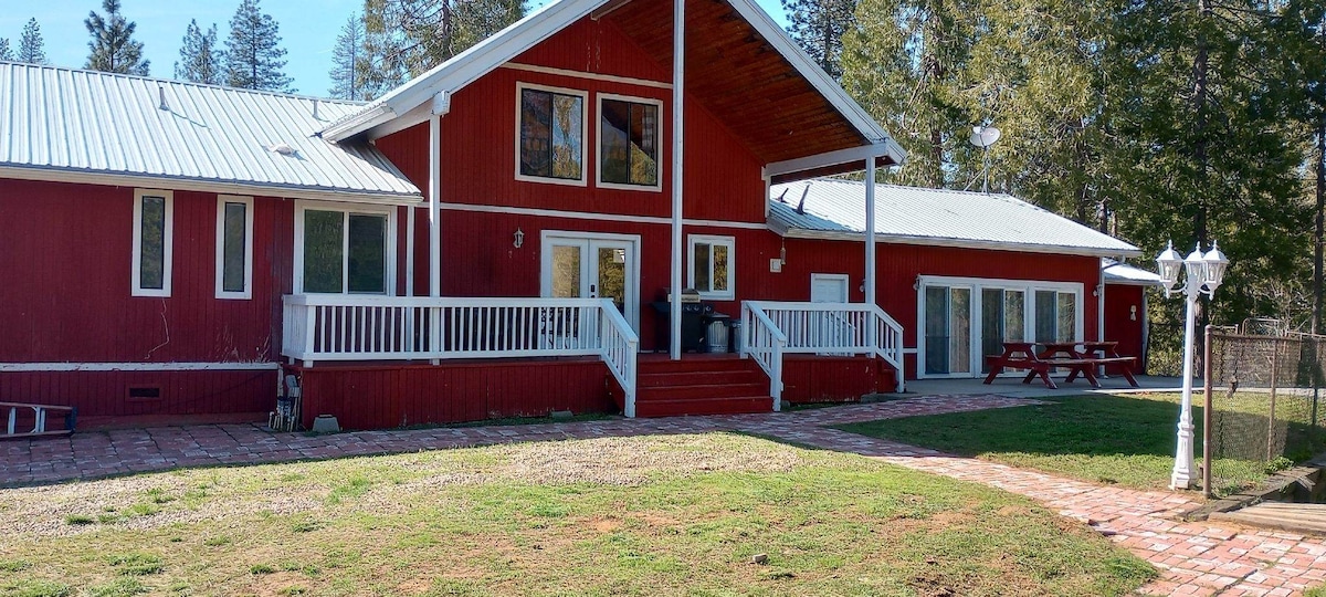 A red cabin is showcased, featuring a spacious front porch and large windows. A brick path leads to the entrance, while green grass fills the yard. A picnic table is positioned nearby, and the surrounding area is bordered by trees.