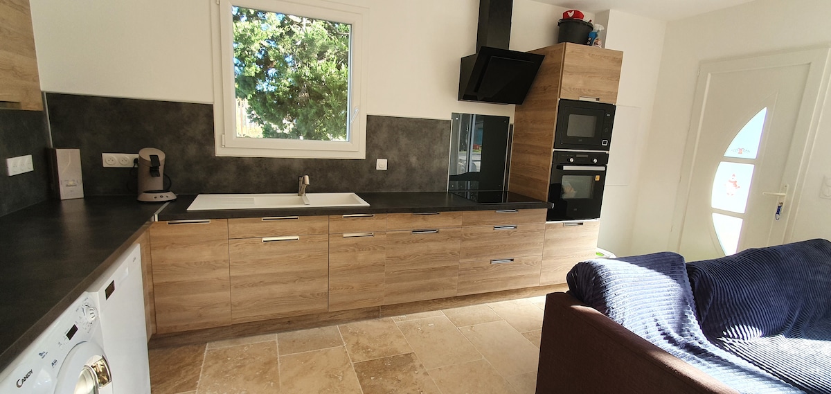 A modern kitchen is equipped with wooden cabinetry and dark countertops. A window allows natural light, illuminating the space. Appliances include a stove, oven, and washing machine, alongside a spacious sink. A cozy sofa is visible in the corner, adding comfort to the area.