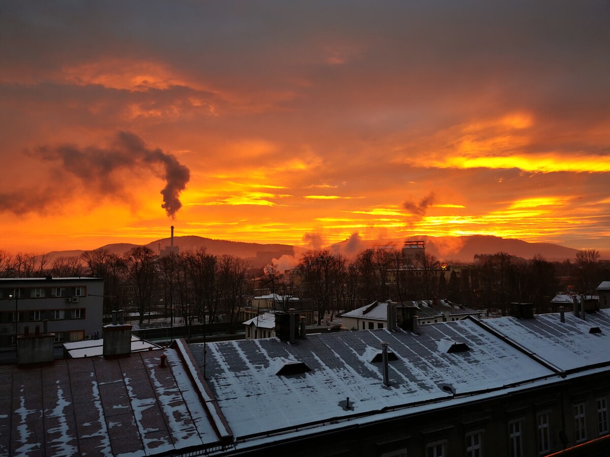 A dramatic sunset casts warm orange and yellow hues across the sky, contrasting with the silhouettes of trees and buildings below. Smoke rises from distant chimneys, indicating an industrial landscape, while rooftops are lightly dusted with snow.