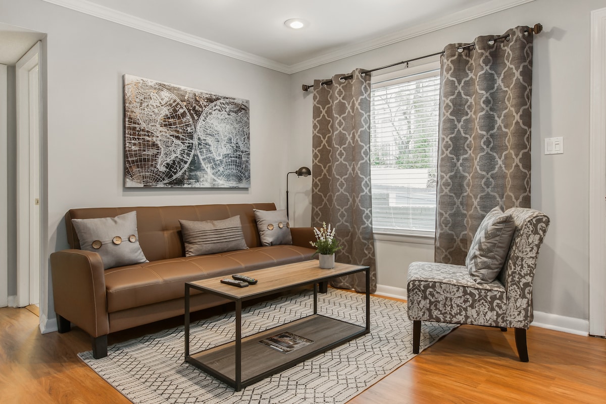 A cozy living room features a modern brown sofa adorned with decorative pillows, complemented by a stylish patterned armchair. Natural light enters through a window dressed with patterned curtains, while a geometric area rug anchors the space. A coffee table displays magazines and a small plant.