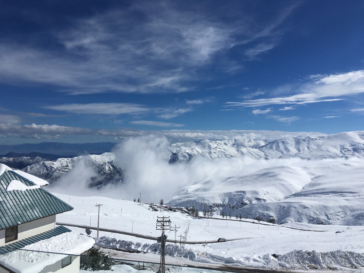 A panoramic view showcases rolling snow-covered mountains under a bright blue sky. Wisps of clouds drift above the landscape, accentuating the vastness of the scene. The foreground features a bit of a building roof, blending naturally with the serene winter setting.