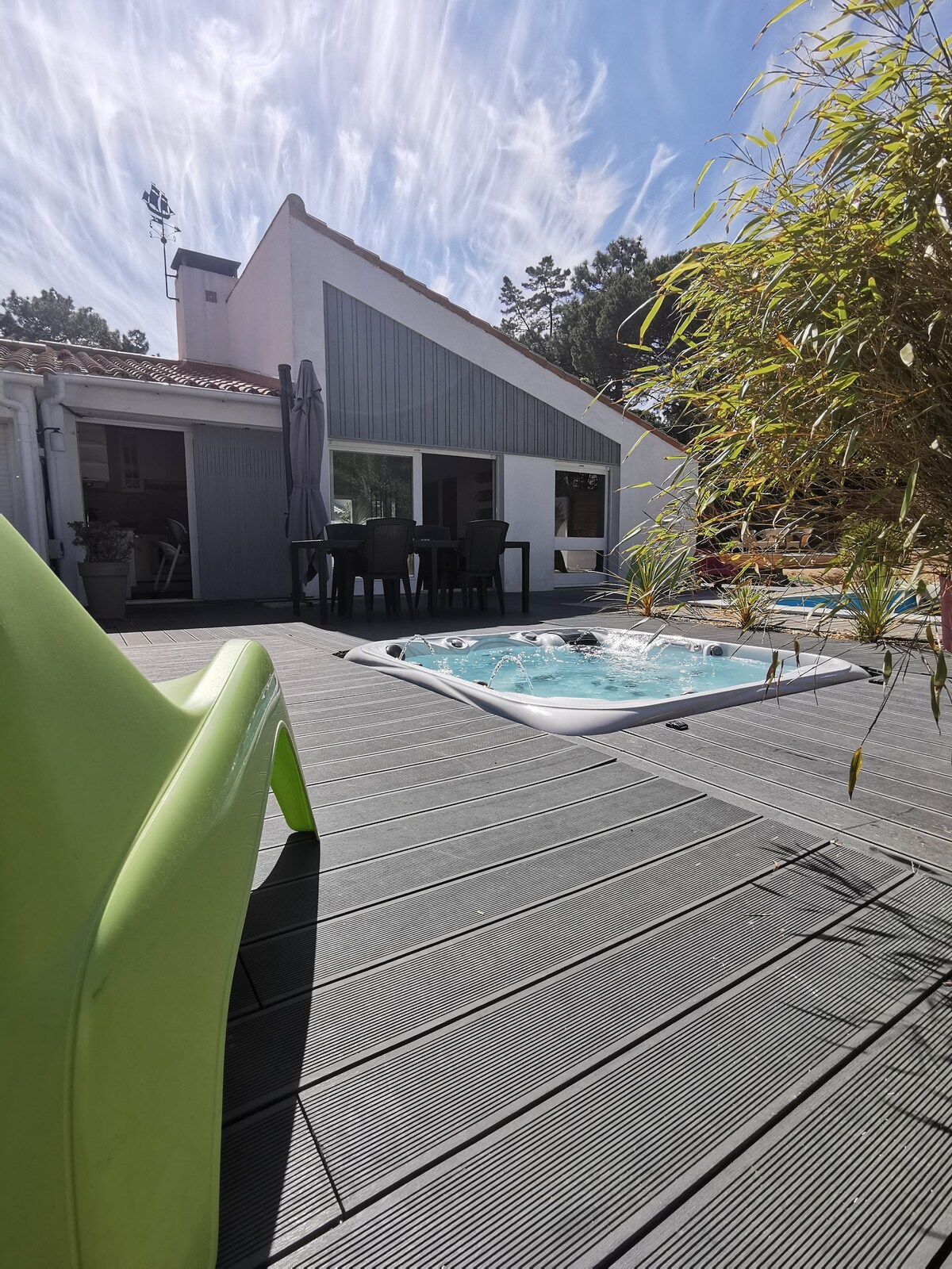 A deck area features a sun lounger and a jacuzzi, with a dining table set for six nearby. The exterior of the house showcases a modern architecture under a clear blue sky. Lush greenery surrounds the space, enhancing the outdoor atmosphere.