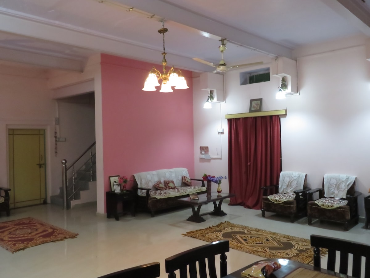 A spacious living area is complemented by soft pink walls and bright ceiling lights. Comfortable seating arrangements, including two sofas and a set of wooden chairs, are arranged around decorative rugs. A curtain frames an entrance leading to another room, contributing to the inviting ambiance.
