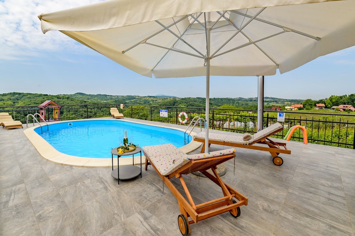 A refreshingly clear pool is framed by a spacious deck, featuring two loungers with patterned cushions under a large umbrella. Lush greenery and distant hills can be seen in the background, creating a serene outdoor setting. A small table with drinks is positioned nearby.