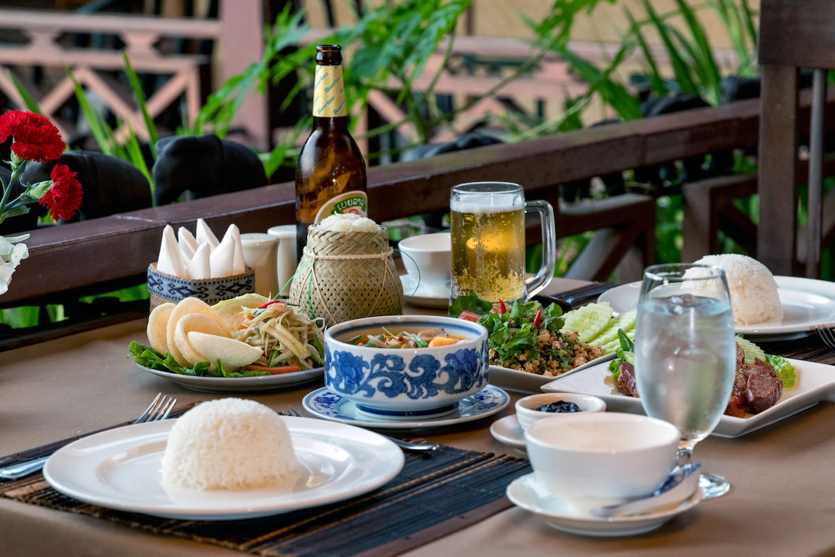 A dining table is set with various dishes, including a bowl of rice, spicy salad, and a traditional Lao meal presented in a decorative bowl. Glasses of water and beer sit alongside napkin holders and condiments, surrounded by greenery for a refreshing touch.