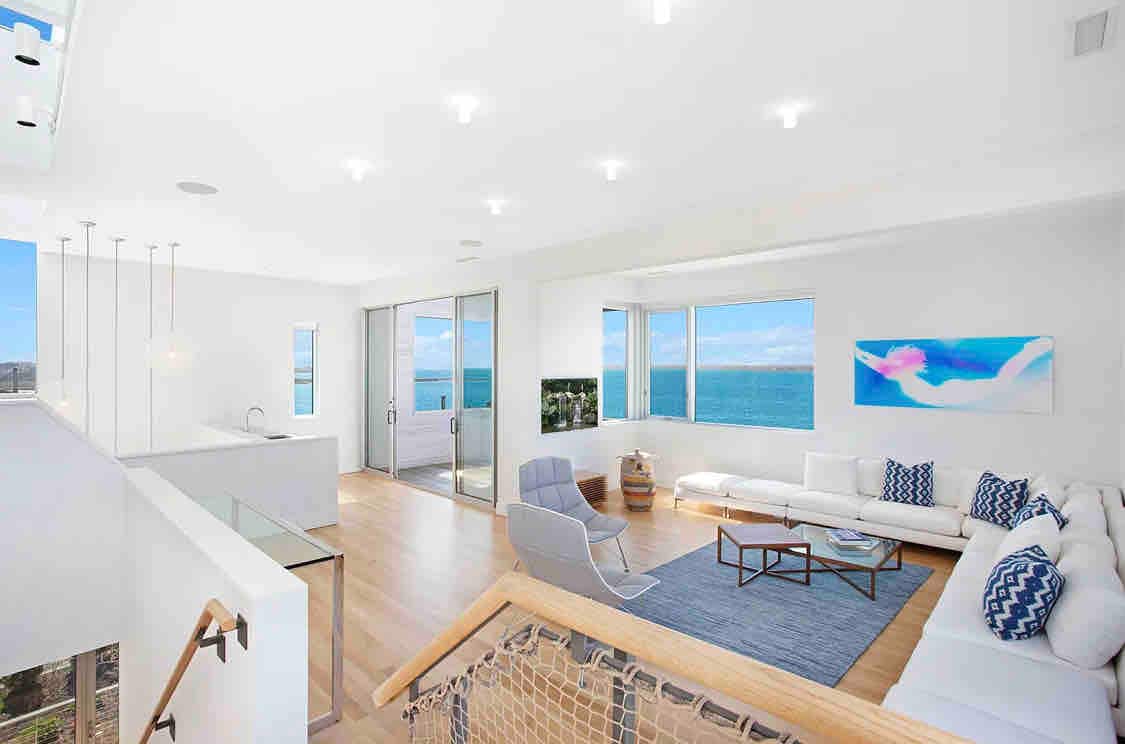 A bright and airy living area showcases floor-to-ceiling windows framing waterfront views. Light hardwood flooring complements the modern furniture, which includes a spacious white sofa adorned with blue and white cushions. An abstract art piece adds color to the wall, contributing to a serene ambiance.