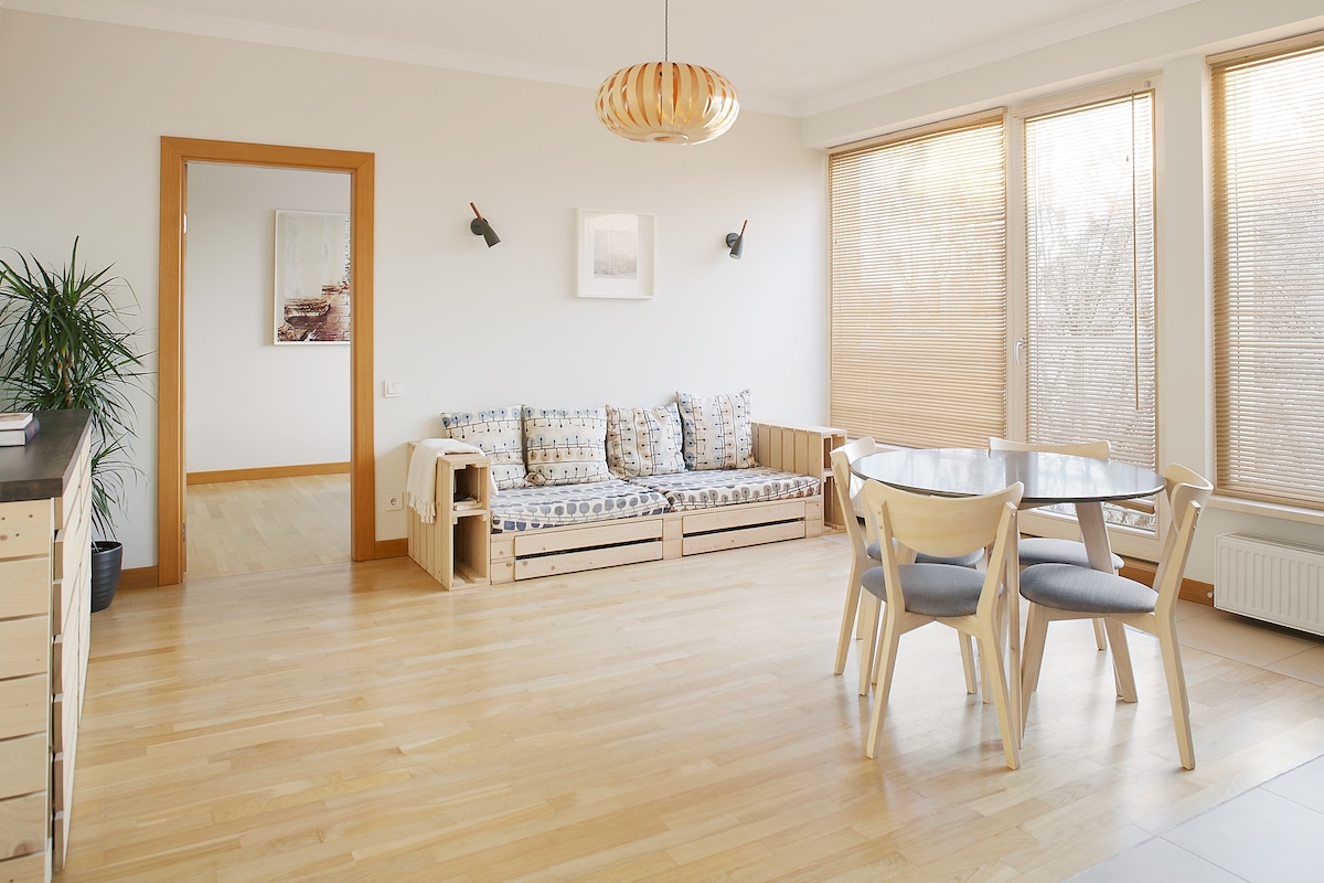 A well-lit living space features large windows with blinds, allowing natural light to fill the room. A comfortable seating area with patterned cushions is visible along one wall, complemented by a round dining table with light chairs. Natural wood flooring enhances the airy atmosphere.