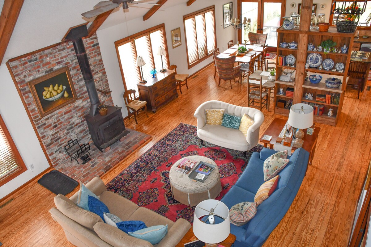 A spacious living area is adorned with wooden beams and warm hardwood flooring. A cozy arrangement of upholstered furniture invites relaxation. A rustic wood stove is situated against a brick accent wall. Shelves display decorative items, and large windows provide ample natural light.