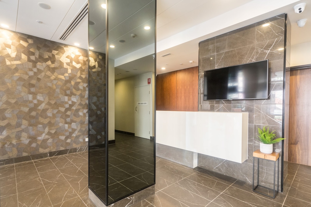 A modern hotel lobby features sleek design elements, including polished stone flooring and a geometric accent wall. A large mirror reflects the space, while a wall-mounted television is positioned above a minimalist reception area. A small plant adds a touch of greenery.