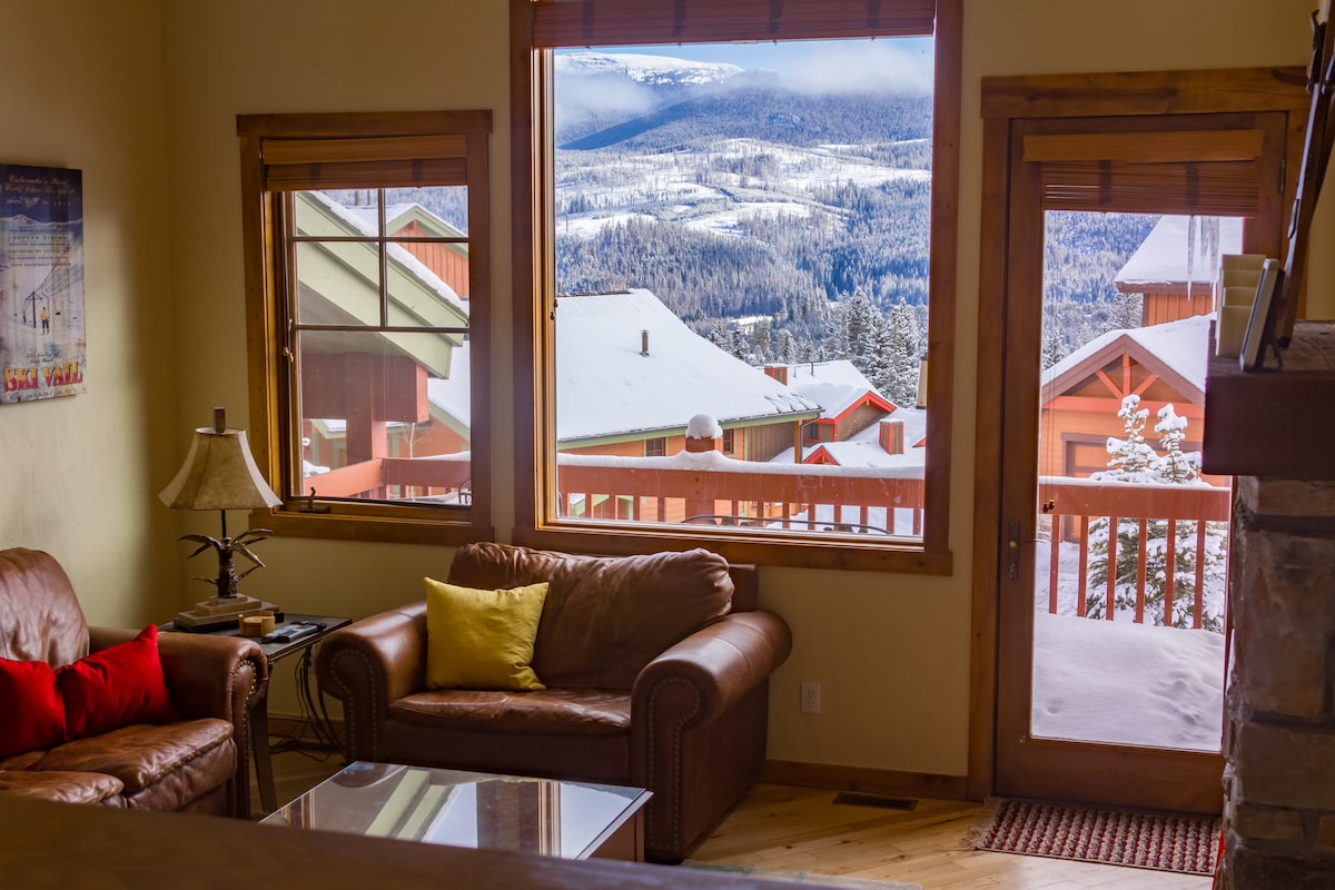 The cozy living area features large windows offering expansive views of snow-covered mountains and rooftops. A leather couch with colorful cushions provides comfortable seating, while natural light brightens the space.