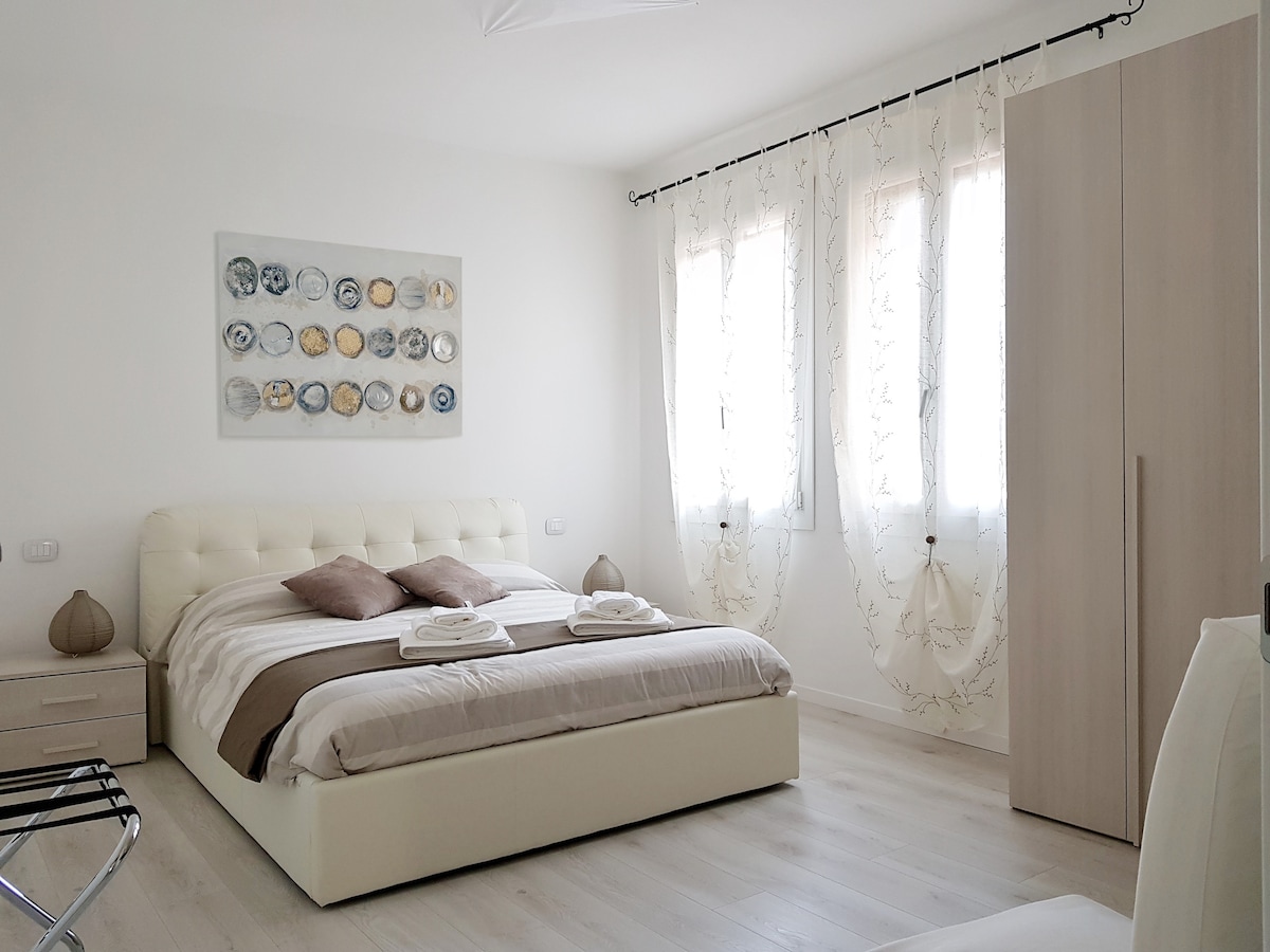 A bright bedroom features a spacious bed dressed in neutral tones and accented by decorative pillows. Two windows with light curtains allow natural light to fill the room. A wardrobe and gentle lighting create a simple yet welcoming space, suited for relaxation.