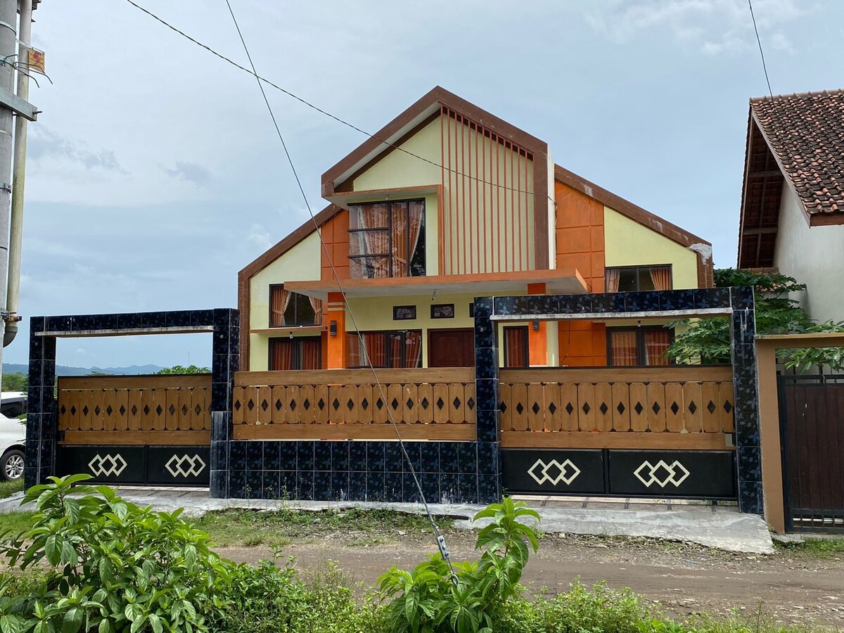 A contemporary two-story house is showcased with bright geometric features in orange and cream. A solid gate with decorative patterns borders the property, with greenery framing the foreground. The windows are large, providing ample light to the interior spaces.