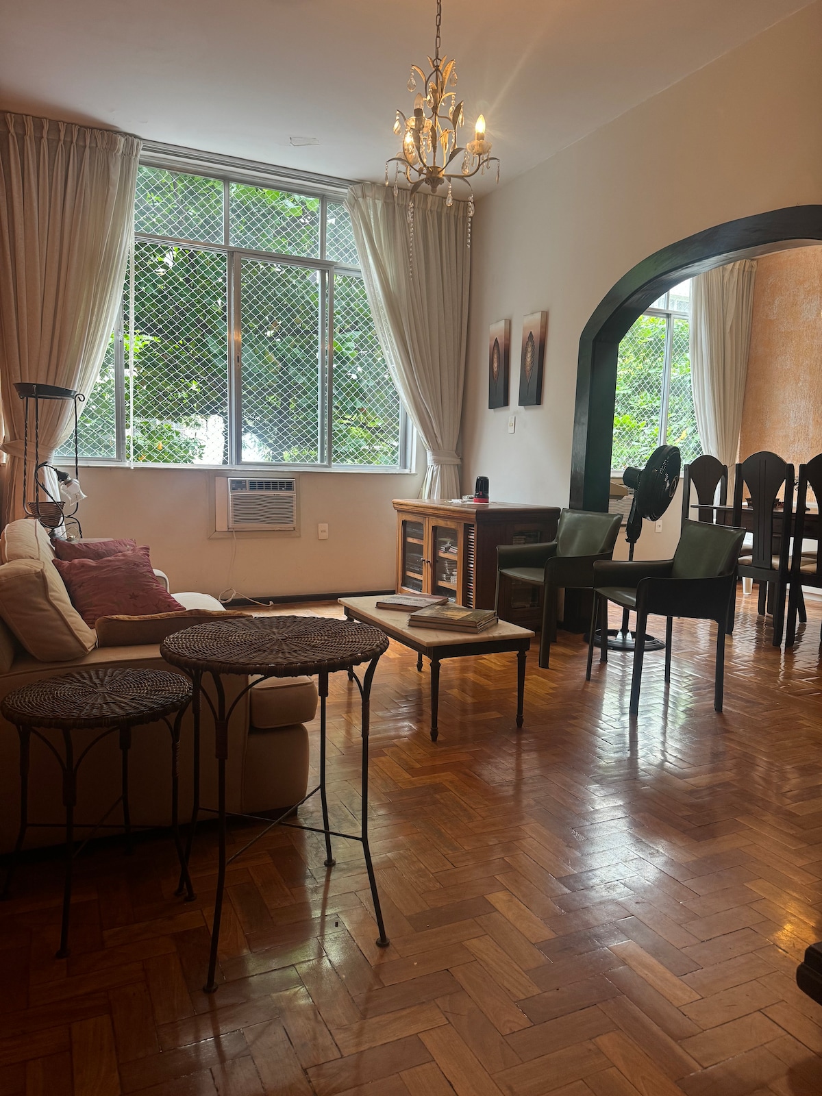 A spacious living area features large windows allowing natural light to enter, accompanied by light-colored curtains. A comfortable sofa is positioned nearby two small decorative tables. A chandelier hangs from the ceiling, while a dining area is visible in the background, furnished with dark chairs.