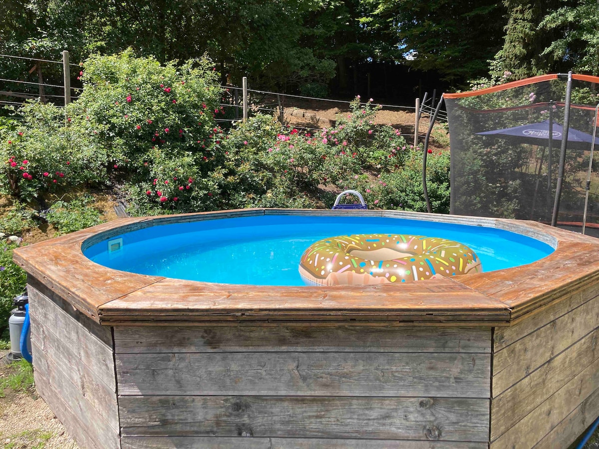 A round above-ground pool is framed by rustic wooden decking, surrounded by lush greenery and blooming flowers. A colorful inflatable ring floats in the clear blue water, inviting relaxation and enjoyment in nature.