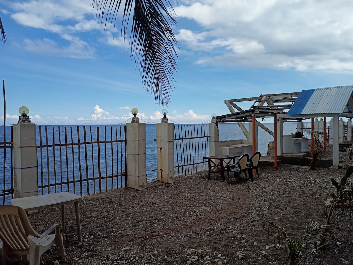 An outdoor area features a gravel surface with a mix of seating arrangements, including a table and chairs. The view of the ocean stretches across the horizon, framed by a fence and complemented by a partially covered structure. Palm trees gently sway in the breeze.