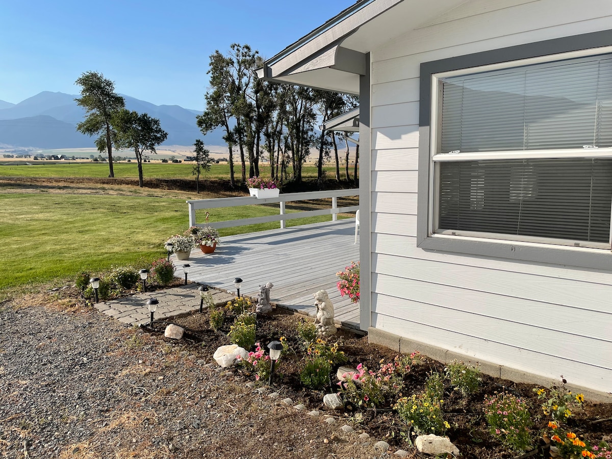 The outdoor area features a well-maintained garden with a variety of colorful flowers and decorative stones. A spacious deck extends from the house, offering views of the surrounding greenery and distant mountains under clear skies.