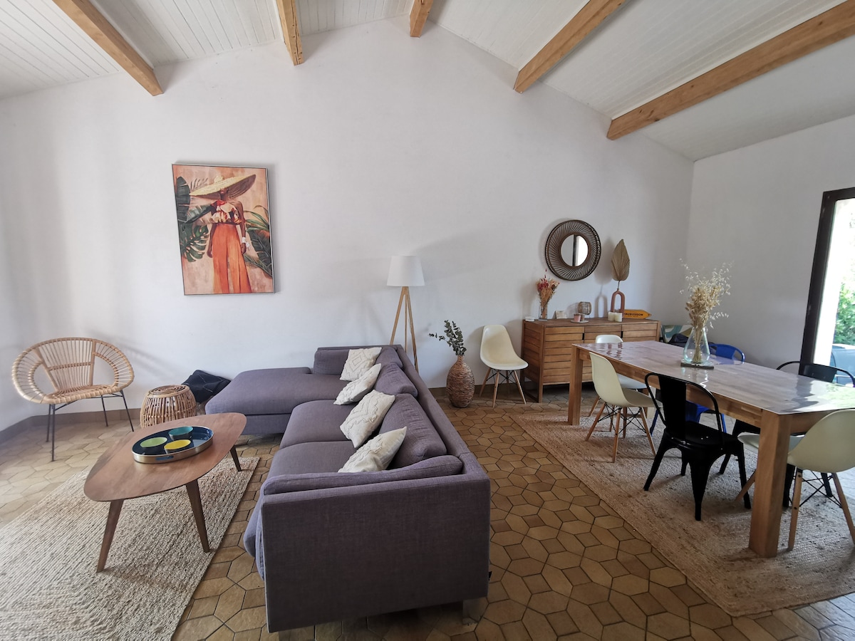A spacious living area is highlighted by wooden beams and warm tones. A comfortable sectional sofa faces a coffee table, while a dining table with mixed chairs occupies the opposite side. A piece of art adorns the wall, and natural light enters through the open doors.