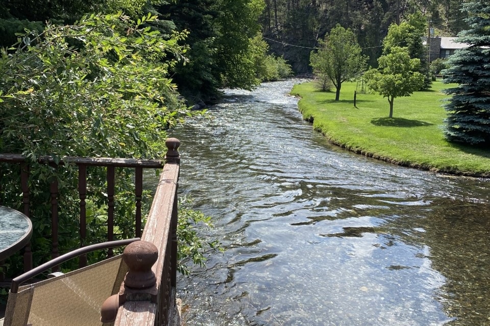 A serene view of Rapid Creek flows gently past the property, surrounded by lush greenery and well-maintained lawns. A comfortable chair and small table are positioned on a wooden deck, inviting relaxation while enjoying the natural setting.