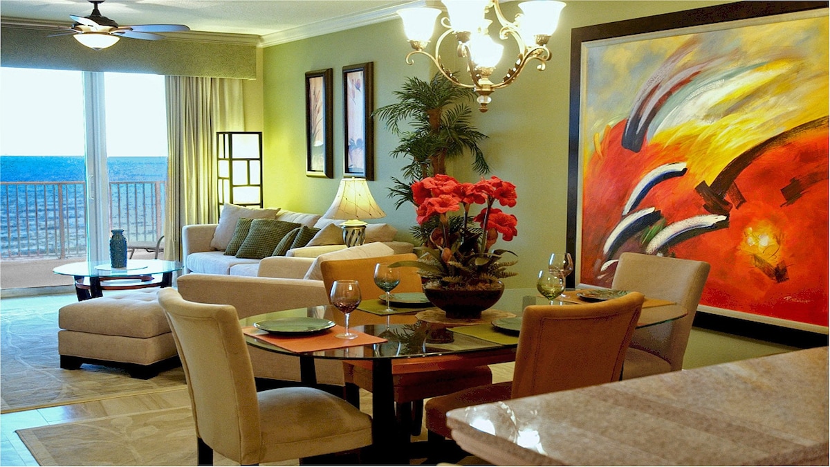 A comfortable living area showcases a light-colored sofa and chairs, with a dining table set for four. A vibrant abstract painting adds a splash of color to the wall. Large windows provide natural light and views of the ocean beyond.
