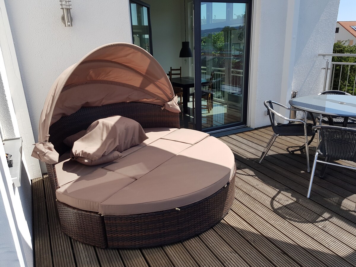 A circular lounge bed with a canopy is positioned on a wooden balcony. Soft tan cushions are neatly arranged on the bed. A small dining table and chairs are visible nearby, with large glass doors leading into the interior space.