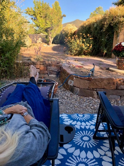 A private patio area is depicted with a textured blue rug laid on gravel. Two lounge chairs are positioned for relaxation, with one chair occupied by a person. Shrubs and flowering plants surround the space, with a glimpse of distant mountains in the background.