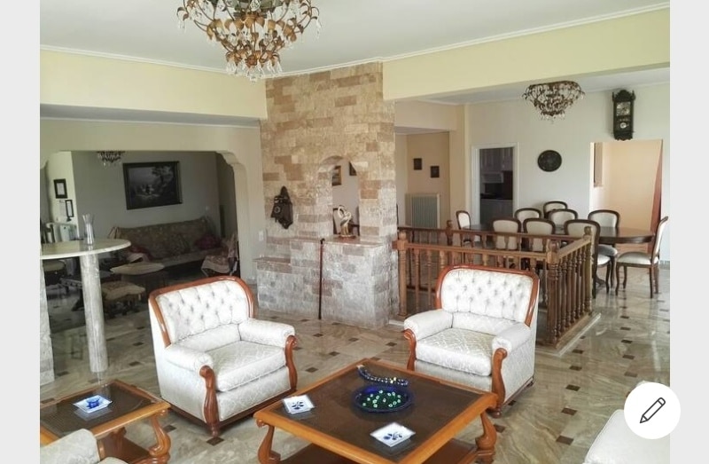 A spacious living area is showcased with elegant furniture, including tufted armchairs and a wooden coffee table. Natural light illuminates the room, highlighting the stone accent wall. The nearby dining area features a wooden table and a series of chairs, enhancing the communal feel of the space.